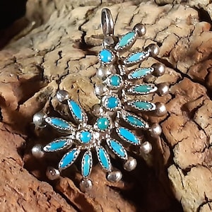 Könnte beinhalten: Silberanhänger mit türkisfarbenen Steinen. Der Anhänger hat ein florales Design mit ovalen und runden türkisfarbenen Steinen, die in Silbermetall gefasst sind. Der Anhänger liegt auf einer Holzoberfläche.