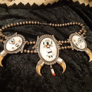 Op de afbeelding: Een zilveren ketting met drie hangers met afbeeldingen van Native American kunst. Elke hanger is omgeven door een zilveren frame en heeft een witte achtergrond met kleurrijke geschilderde figuren. De ketting is versierd met zilveren kralen en klauwvormige accenten.