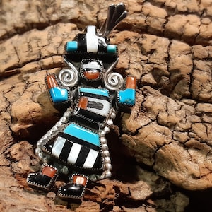 Könnte beinhalten: Ein silberner Anhänger mit einer stilisierten Figur mit Türkis-, Schwarz-, Weiß- und Roteinlegearbeiten. Die Figur trägt einen Kopfschmuck und hat ein geometrisches Design.