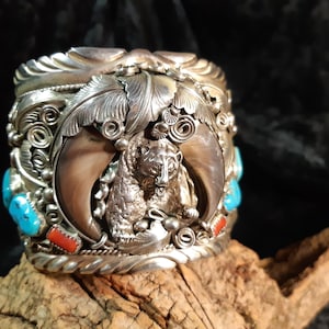 Può includere: Bracciale a polsino in argento con un design di orso e artiglio. Il bracciale presenta accenti di pietre turchesi e rosse, insieme a intricati dettagli di foglie e volute. L'orso è centrato, incorniciato da grandi artigli.