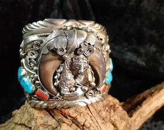 May include: Silver cuff bracelet with a bear and claw design. The bracelet features turquoise and red stone accents, along with intricate leaf and swirl details. The bear is centered, framed by large claws.