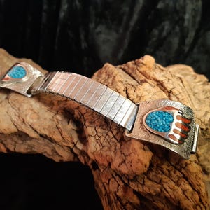 Puede incluir: Correa de reloj plateada con diseño de pata de oso turquesa y rojo. La correa del reloj tiene una banda de metal rectangular y extensible. El diseño de la pata de oso está incrustado con pequeñas piedras turquesas y detalles rojos.