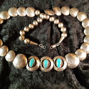 Peut inclure: Collier argenté composé de grosses perles gravées en forme de disque et de petites perles rondes. Le collier comprend des pierres de couleur turquoise serties dans des lunettes en argent. Le collier est présenté sur une surface sombre et texturée.