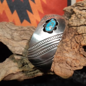 Op de afbeelding: Een zilveren armband met een turquoise steen in een zilveren zetting. De armband heeft een decoratief ontwerp van lijnen en spiralen.
