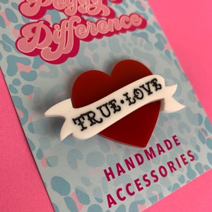 May include: Red heart-shaped pin with a white banner that reads "TRUE LOVE" on a blue and white leopard print background. The pin is handmade and says "Peggy's Difference" and "Handmade Accessories" on the card.