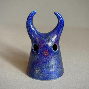 May include: A small, cone-shaped ceramic sculpture with a glossy, deep blue glaze featuring purple and black mottled accents. The object has two prominent, curved horns extending from the top and two circular eye holes with a small central opening.