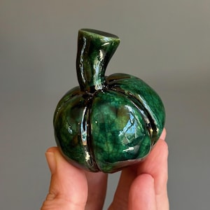 May include: A small, rounded ceramic sculpture shaped like a pumpkin with a glossy, deep emerald green glaze. The surface features a distinct crackle pattern throughout the dark green finish, with a prominent, curved stem extending from the top.