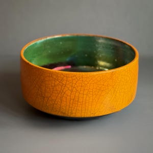 May include: A handmade ceramic bowl. The exterior is a vibrant orange with a cracked, textured finish. The interior is green with a glossy finish and hints of pink and other colours. The bowl is suitable for serving food.