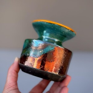May include: A small ceramic vessel with a wide, flared rim and a cylindrical body. The piece features a vibrant glaze transition from a bright orange rim to a deep teal midsection, ending in a dark, textured brown base with a glossy finish.