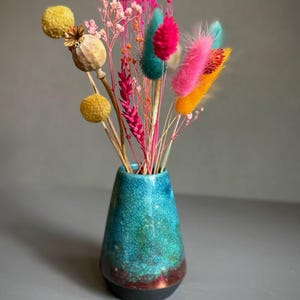 Può includere: Un vaso in ceramica turchese craquelé contiene una vivace composizione di fiori secchi. Il bouquet include accenti gialli, rosa e turchese. Il vaso ha una base marrone scuro e una forma svasata, creando un contrasto sorprendente.