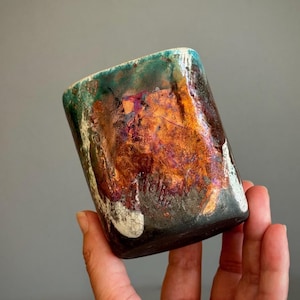 May include: A cylindrical ceramic cup featuring a multi-coloured glaze with prominent metallic copper, bronze, and deep purple tones. The upper rim displays a vibrant teal green accent, while the base transitions into a dark, charcoal-coloured finish.