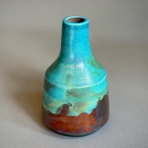 Könnte beinhalten: Eine Keramikvase mit engem Hals und breitem Boden. Der obere Teil ist mit einer leuchtend türkisfarbenen Glasur mit sichtbarem Rissmuster versehen, während der untere Bereich in ein tiefes, erdiges Braun und einen kupferfarbenen Metallic-Ton übergeht.