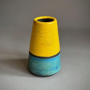Könnte beinhalten: Eine Keramikvase in konischer Zylinderform mit einem leuchtend gelben oberen Teil und einem türkisfarbenen unteren Teil. Beide farbigen Bereiche weisen eine sichtbare Craquelé-Glasur auf, die durch ein schmales schwarzes Band getrennt ist.