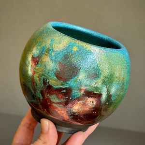 May include: A handcrafted ceramic bowl with a unique, textured glaze. The bowl features a blend of teal, green, and reddish-brown hues, with a dark base. The interior is a vibrant teal color.
