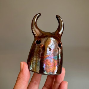May include: A small, hand-sculpted ceramic figurine with two prominent, curved horns extending from the top. The surface features a glossy, metallic brown glaze with iridescent, rainbow-like highlights and two circular cutouts for eyes.