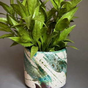 May include: A decorative ceramic plant pot with a white base and teal and brown brushstroke design. The pot is filled with bright green leafy plants. The pot has a textured, crackled finish. The pot is a stylish home decor item.