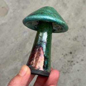 May include: A ceramic mushroom sculpture featuring a glossy, crackled emerald green glaze on the cap and stem. The stem transitions into a metallic, iridescent copper and dark purple finish near the base, which is a matte, unglazed black texture.