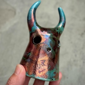 Può includere: Una scultura in ceramica con due corna ricurve verso l'alto e una finitura liscia e lucida. La superficie presenta una miscela marmorizzata di toni rame metallizzato, bronzo e verde ottanio, con due aperture circolari visibili sul davanti.