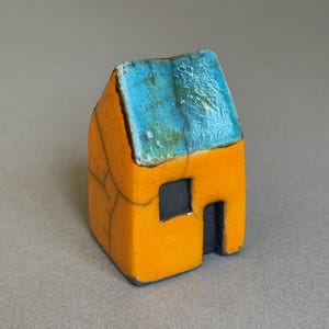 May include: A small ceramic house figurine featuring a bright yellow exterior with a visible crackle glaze pattern. The roof is finished in a textured, glossy turquoise blue glaze. It includes a single dark grey square window and a dark grey doorway.