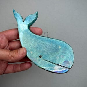 May include: A ceramic whale ornament featuring a glossy turquoise glaze with a fine crackle finish. The whale has a small metal loop attached to the top for hanging, a single dark eye, and a horizontal line detail representing the mouth.