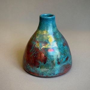 May include: A small ceramic vase with a wide base and narrow neck, featuring a glossy turquoise glaze with a fine crackle pattern. The surface displays irregular patches of deep red, metallic gold, and iridescent highlights across the body.