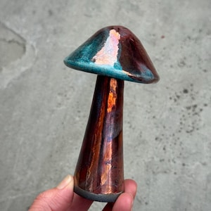 Puede incluir: Una escultura de hongo de cerámica con un esmalte brillante e iridiscente. El sombrero presenta una mezcla de tonos azul verdoso profundo y cobre metálico, mientras que el tallo alto y delgado exhibe un acabado reflectante en bronce y marrón.