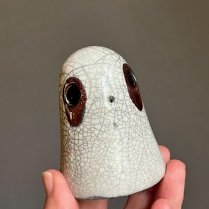 May include: A ceramic ghost figurine with a white crackle glaze. The ghost has two large, dark brown eye sockets with black pupils. The figurine is held in a hand against a neutral background.
