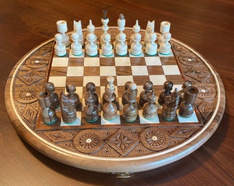 May include: A wooden chess set with intricate carvings on the board and pieces. The board is round and has a hinged lid. The pieces are made of wood and have a light brown and dark brown finish.