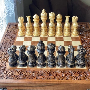 Op de afbeelding: Een houten schaakbord met ingewikkelde houtsnijwerk op de lijst. Het bord is open en toont een set gouden en zwarte schaakstukken. De stukken zijn gerangschikt in een beginpositie voor een schaakspel.