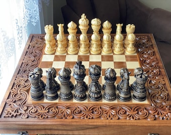 Può includere: Una scacchiera di legno con intagli intricati sul telaio. La scacchiera è aperta e mostra un set di pezzi degli scacchi dorati e neri. I pezzi sono disposti in una posizione di partenza per una partita a scacchi.
