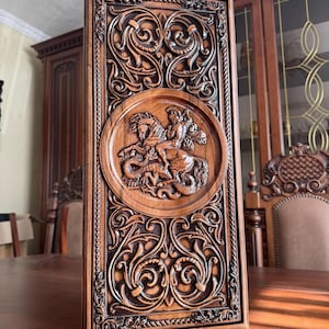 May include: A rectangular, wooden backgammon board with detailed, carved floral and scrollwork patterns. A central circular relief shows a figure on horseback slaying a dragon. The board is a rich, warm brown colour.