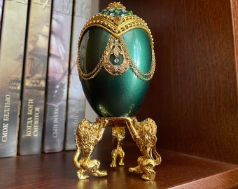 May include: A green jeweled egg with gold accents sits on a gold stand with three lion feet. The egg is decorated with gold chains and a jeweled crown.