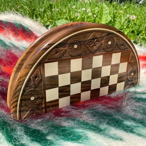 Puede incluir: Un tablero de ajedrez de madera en forma de semicírculo con intrincados tallados alrededor del borde. El tablero está hecho de madera oscura con una incrustación de madera clara para las casillas. El tablero está sentado sobre una manta peluda multicolor.