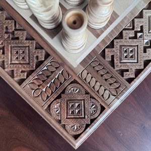May include: Close-up of a wooden chessboard with intricate carvings. The board features a geometric pattern with leaves and squares. The chess pieces are made of light wood and are arranged in the center of the board.