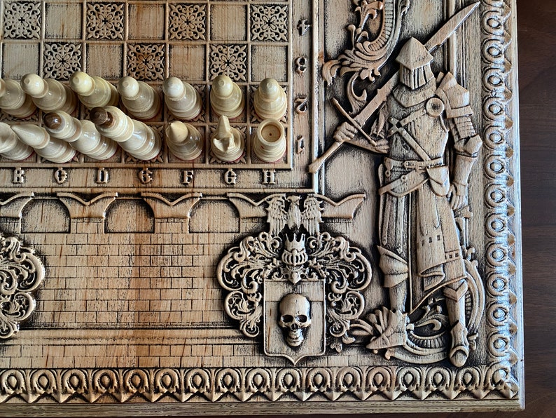 Op de afbeelding: Een houten schaakbord met ingewikkelde houtsnijwerk van een ridder, een wapenschild en een schedel. Het bord heeft een lichtbruine afwerking en is versierd met een rand van kleine cirkels. De schaakstukken zijn wit en zijn op het bord gerangschikt.