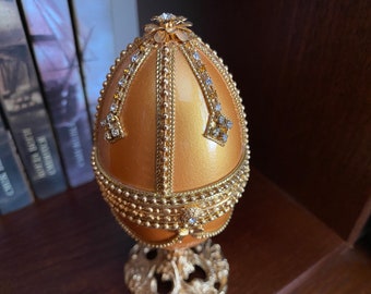 May include: A gold egg-shaped music box with a decorative design of gold beads and rhinestones. The music box is on a gold stand with intricate details.