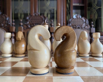 May include: Two wooden chess pieces, a white knight and a brown knight, stand facing each other on a chessboard. The knights are carved in a unique style with a curved, S-shaped neck.