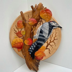 Puede incluir: Una pieza de arte de pared de madera tallada que representa un pájaro carpintero posado en una rama con hojas de otoño. El pájaro está pintado con detalles realistas con plumas blancas y negras y una cresta roja. Las hojas están pintadas en tonos de naranja, rojo y amarillo.