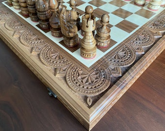 May include: A wooden chessboard with intricate carvings around the edge. The board is closed and the pieces are in place. The board is made of dark wood and has a light wood inlay.