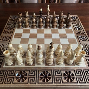 Könnte beinhalten: Ein Schachspiel aus Holz mit einem Brett aus Nussbaum und Ahorn. Das Brett hat einen dekorativen Rand mit einem griechischen Schlüsselmuster. Die Figuren sind aus Holz und befinden sich in der Ausgangsposition.