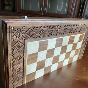 Puede incluir: Un tablero de ajedrez de madera con intrincados tallados alrededor del borde. El tablero está cerrado y tiene un acabado marrón claro. El tablero de ajedrez está hecho de madera y tiene un diseño tradicional.