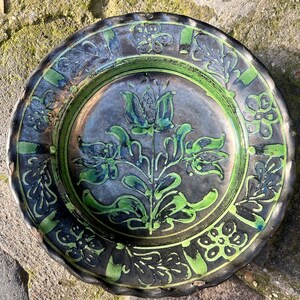 Könnte beinhalten: Ein runder Keramikteller mit einem grünen und schwarzen Blumendesign. Der Teller hat einen gewellten Rand und ist mit einer stilisierten Blume in der Mitte verziert.