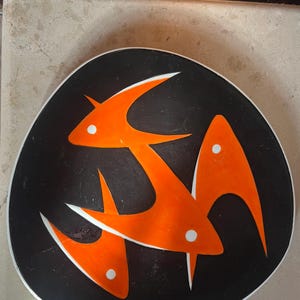 Pode incluir: Um prato de cerâmica preto com um design estilizado de três peixes laranja com detalhes brancos.