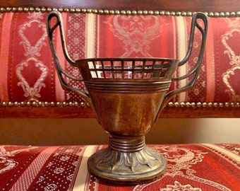 Op de afbeelding: Een zilverkleurige decoratieve metalen beker met twee gebogen handvatten. De beker heeft een roosterachtige rand en een gedetailleerde basis met een bloemmotief. De beker wordt gepresenteerd op een rood-wit oppervlak met patronen.