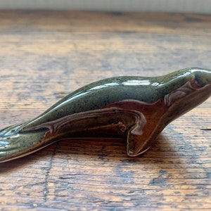 May include: A ceramic seal figurine with a green and brown glaze. The seal is lying on its belly with its head turned to the side.