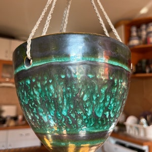 Puede incluir: Un macetero colgante de cerámica verde y negro con un asa de cuerda. El macetero tiene un esmalte moteado y un borde verde oscuro.