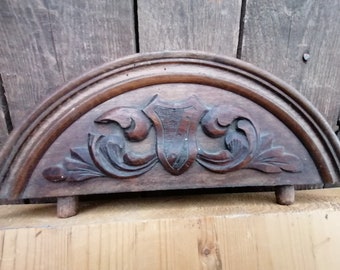 Op de afbeelding: Een gegraveerde houten halfronde boog met een schild en een bloemmotief. De boog is donkerbruin en heeft een verweerde afwerking.