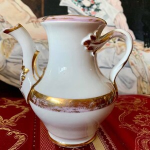 Könnte beinhalten: Eine weiße Porzellan-Teekanne mit rosafarbenem Rand und goldenen Akzenten. Die Teekanne hat ein zartes, verziertes Design und einen geschwungenen Griff.
