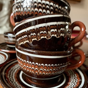 May include: A set of four brown ceramic coffee cups with white and brown swirl designs. The cups are stacked on top of each other and are sitting on a brown saucer with a similar design.