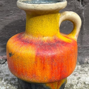 Op de afbeelding: Een kleine keramische vaas met een handvat. De vaas heeft een gele bovenkant, overgaand in oranje en rood in het midden, en een zwarte basis. Het handvat en de rand zijn crèmekleurig. Ongeveer 15 cm hoog.